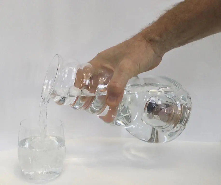 Man sieht eine männliche Hand, die aus dem Acala Cadus Krug mit Edelsteinfach Wasser in ein Glas kippt. Weißer Hintergrund.