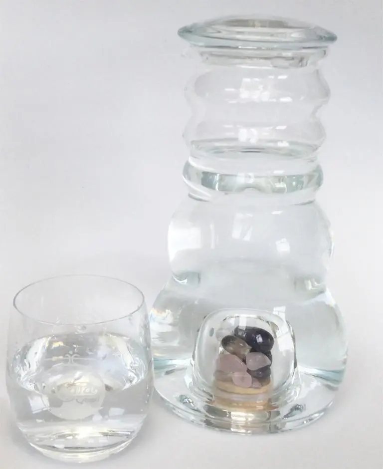 Zu sehen ist eine mit Wasser gefüllte Glaskaraffe mit flachem Glasdeckel und ein mit Wasser gefülltes Trinkglas. Die Karaffe hat im unteren Teil ein separates Fach mit Edelsteinen. Das Bild zeigt den Cadus Krug und das Arlando Glas von Acala.