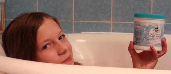 Zu sehen ist ein Mädchen mit braunen Haaren, was in einer Badewanne liegt. Man sieht den oberen Rand der Wanne, den Kopf und die Hand des Mädchens. Sie schaut in die Kamera und hält eine Dose Basenbad 700g in der Hand.