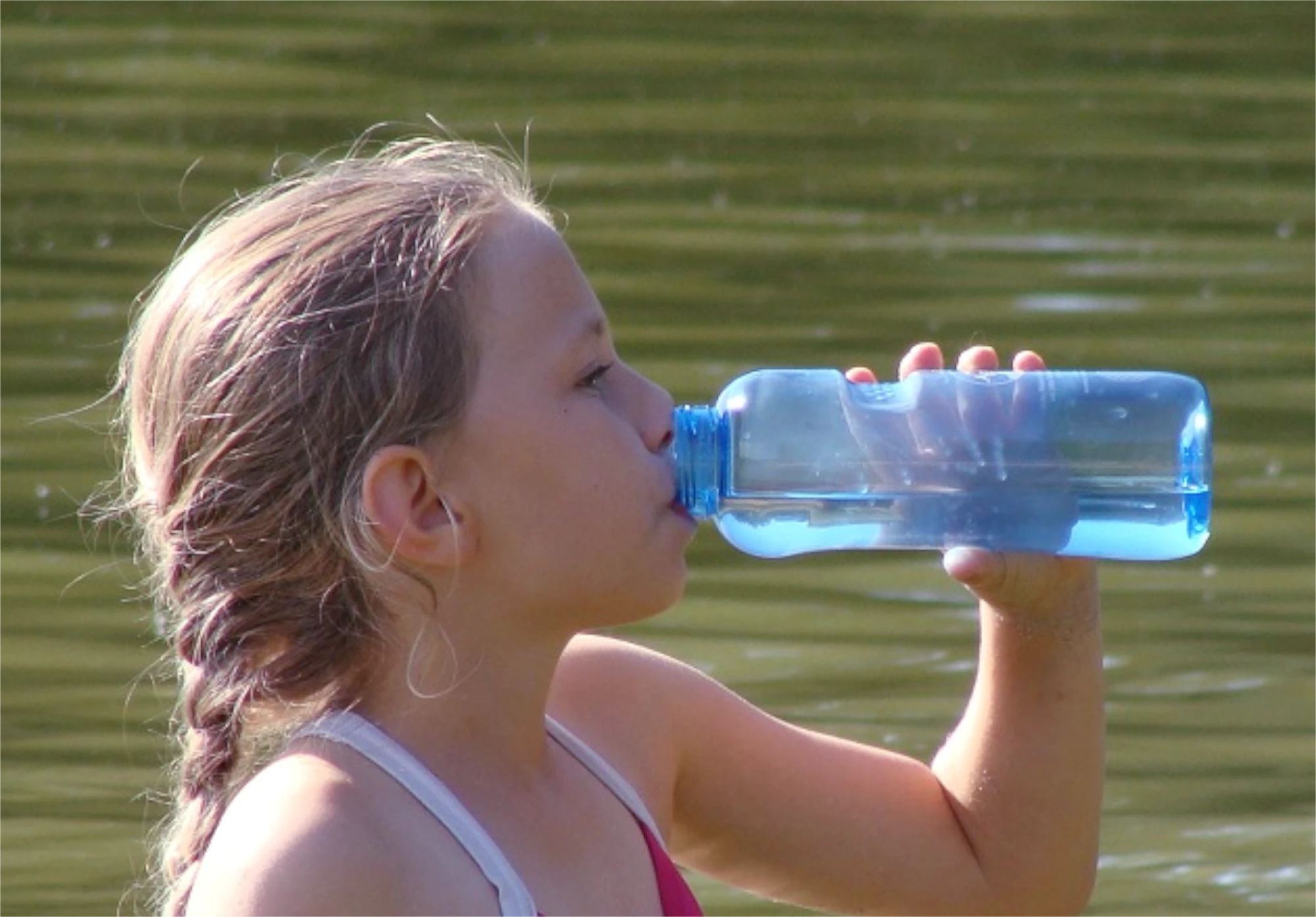 Kleines Mädchen mit geflochtenem Pferdeschwanz am See trinkt Wasser aus einer blauen Tritan Trinkflasche von Acala.