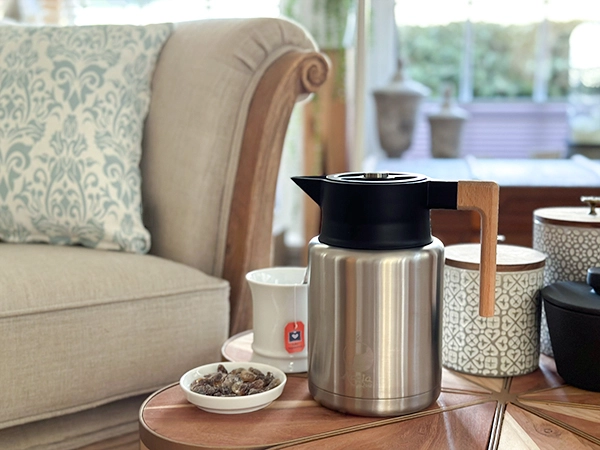 Thermoskanne 1,5L edel & nachhaltig Zu sehen ist eine Thermoskanne in einem Wohnzimmer auf einem kleinen Holztisch vor einem hellen Sofa mit einem Kissen darauf. Es steht eine Tasse und Kandis Zucker in einer kleinen Schale neben der Kanne. Das Bild zeigt die Acala Thermoskanne.