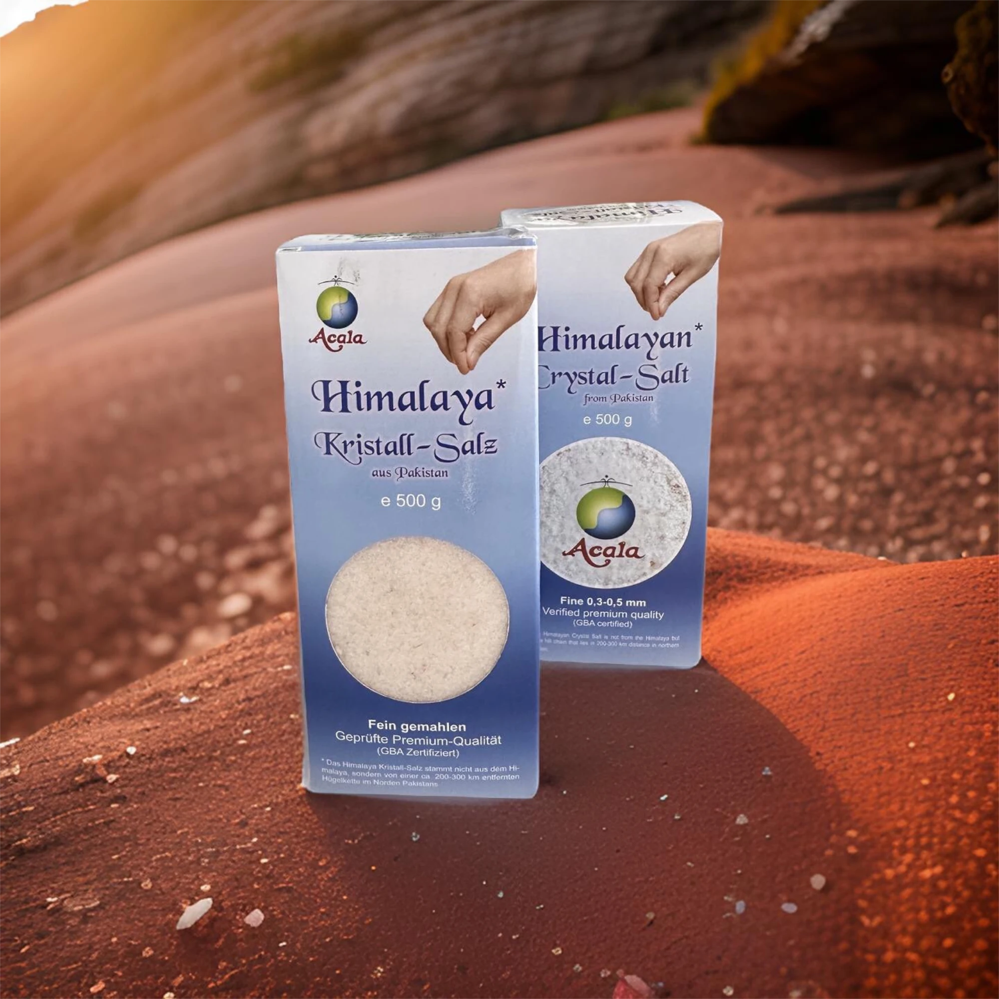 Zu sehen ist ein helles Holzbrett. Auf diesem steht eine grüne Papiertüte mit Kristallsalz von Acala. Des Weiteren sieht man eine mit diesem Salz gefüllte Gewürzmühle, gebratenes Lachsfilet und verschiedenes Gemüse, wie Tomaten, Paprika und Gurken.