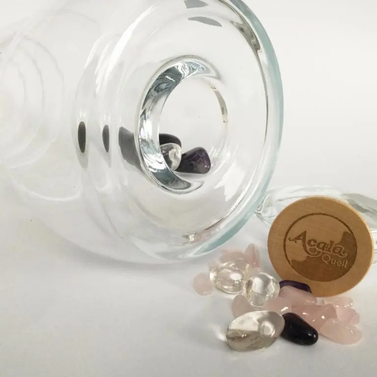 Zu sehen ist der Boden einer Glaskaraffe. Man das Loch des separaten Edelsteinfachs und davor liegen verschiedene Edelsteine und der Holzverschluss mit Acala Logo für das Edelsteinfach. Das Bild zeigt den Cadus Krug mit Edelsteinfach von Acala.