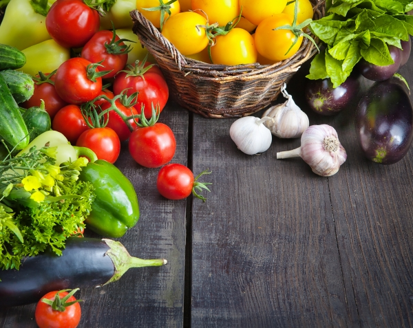 Zu sehen ist verschiedenes, frisches Gemüse auf einem dunkeln Holztisch. Tomaten, Knoblauch, Paprika, Auberginen, Gurken und Salat. 