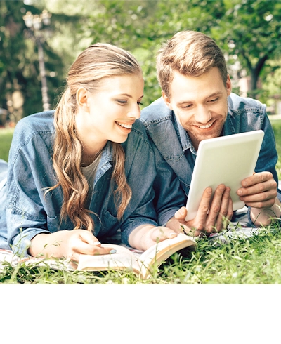Zu sehen ist eine junge Frau mit langen blonden Haaren und ein junger Mann mit kurzen blonden Haaren. Beide tragen ein blaues Jeanshemd, liegen im grünen Gras und schauen lächelnd auf ein Tablet.