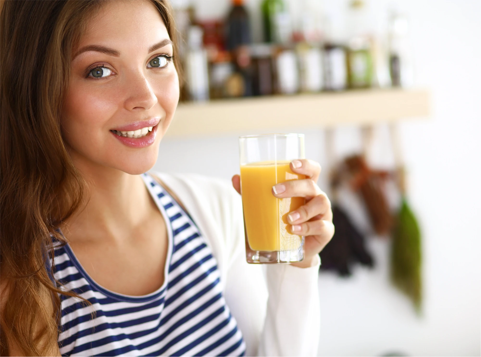 Junge Frau mit langen braunen Haaren in heller Küche schaut lächelnd in die Kamera und hält ein Glas Orangensaft in der Hand.