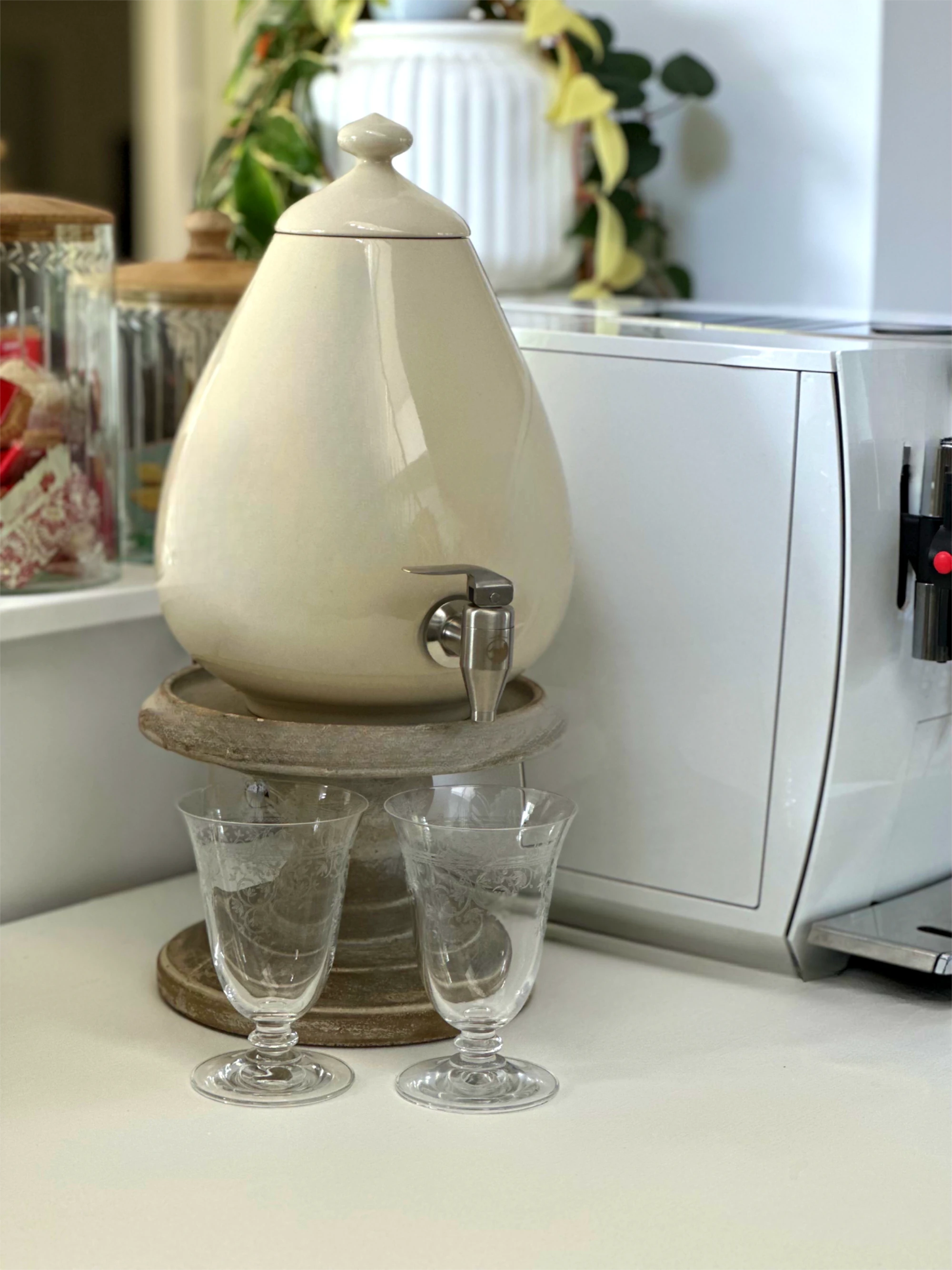 Zu sehen ist ein 5L Porzellan Ei aus Handarbeit von Acala. Es steht auf einem Holzsockel, darunter stehen 2 Gläser. Das Ei ist beige und es ist Edelstahl Wasserhahn daran. Neben dem Ei sieht man einen Kaffeevollautomaten.