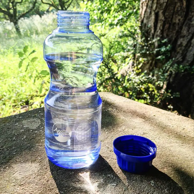 Zu sehen ist eine offene, mit Wasser gefüllte blaue Tritan Trinkflasche. Sie steht auf einem Stein, im Hintergrund sieht man einen Baumstamm und eine grüne Wiese. Neben der Flasche liegt der dunkelblaue Schraubdeckel. Man sieht die EVI Flasche von Acala.
