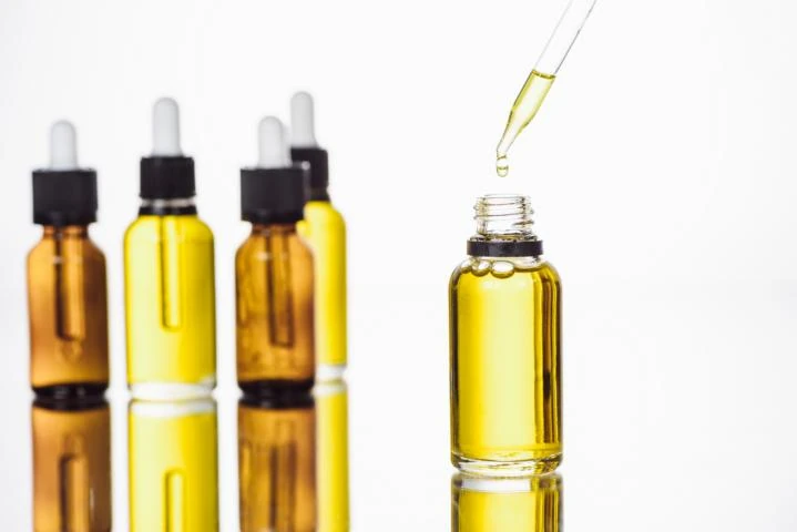 Vier kleine Flaschen gefüllt mit gelber und brauner Flüssigkeit und Pipetten vor weißem Hintergrund. Im Vordergrund eine offene kleine Flasche mit gelber Flüssigkeit, ein Tropfen wird aus der Pipette in die Flasche getropft.