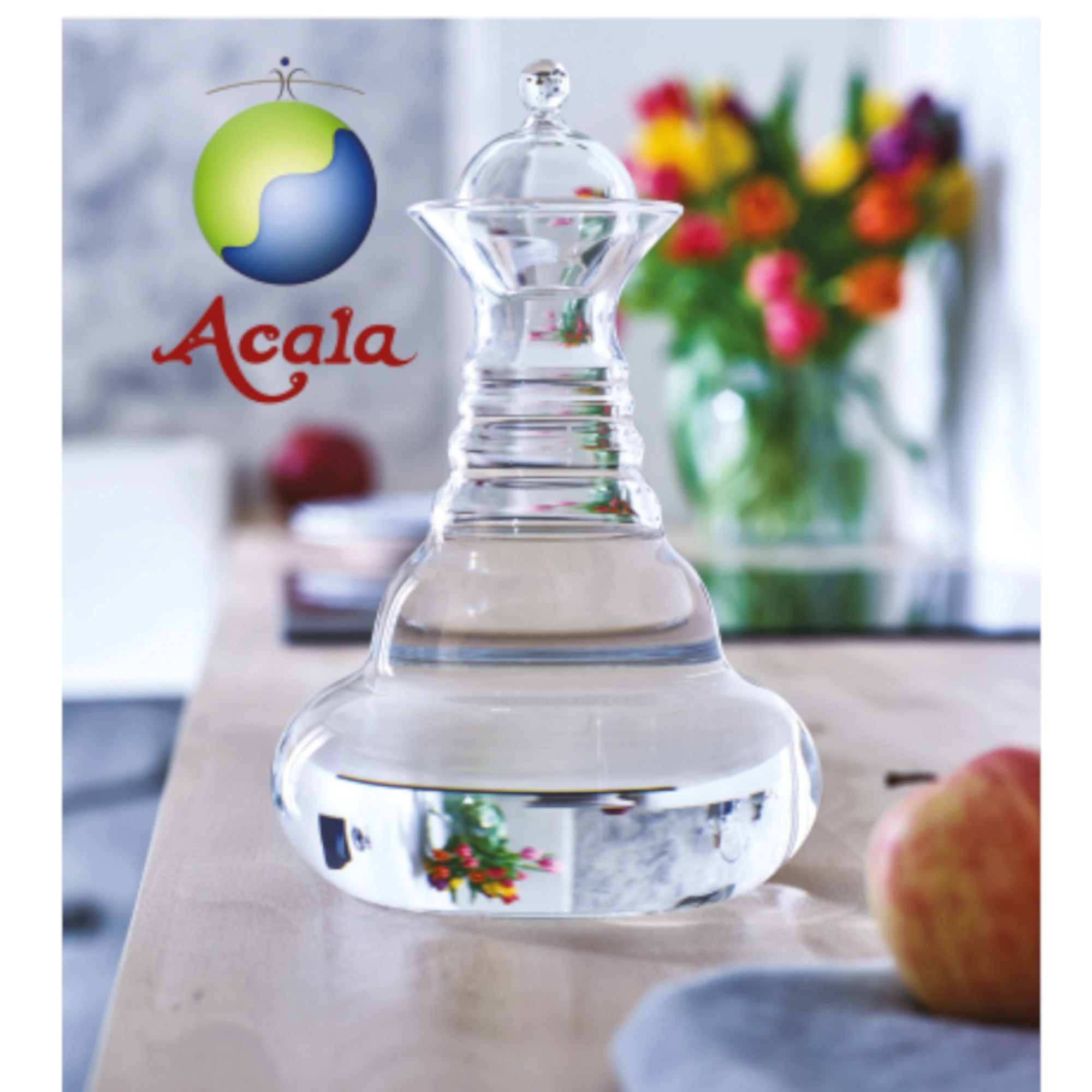 Alladin Karaffe Gold mit Glasdeckel und Blume des Lebens 1,3 Liter Zu sehen ist die Alladin Glas Karaffe von Acala mit Glasdeckel. Sie steht auf einem Tisch, man sieht Obst und einen bunten Blumenstrauß. Links neben der Karaffe sieht man das Acala Logo.