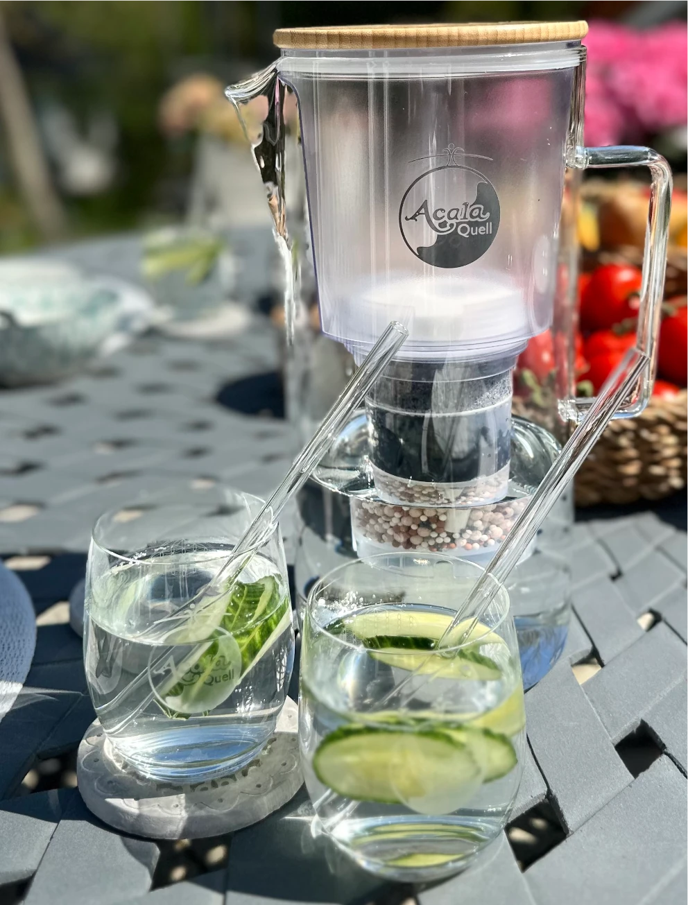 Zu sehen ist der Glas Kannenfilter Silizia von Acala auf einem Gartentisch im Sommer. Davor stehen 2 Gläser gefüllt mit Wasser, Gurken und Glasstrohhalmen. Im Hintergrund sieht man verschwommen eine Schale mit Gemüse.