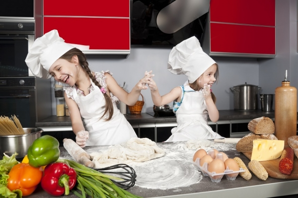 Zu sehen sind zwei kleine Mädchen, etwa 6 Jahre alt, mit weißer Schürze und großen weißen Kochmützen auf dem Kopf in einer Küche mit roten Fronten. Vor Ihnen liegt Teig in Mehl auf der Arbeitsplatte. Sie bewerfen sich gegenseitig mit Mehl und lachen.