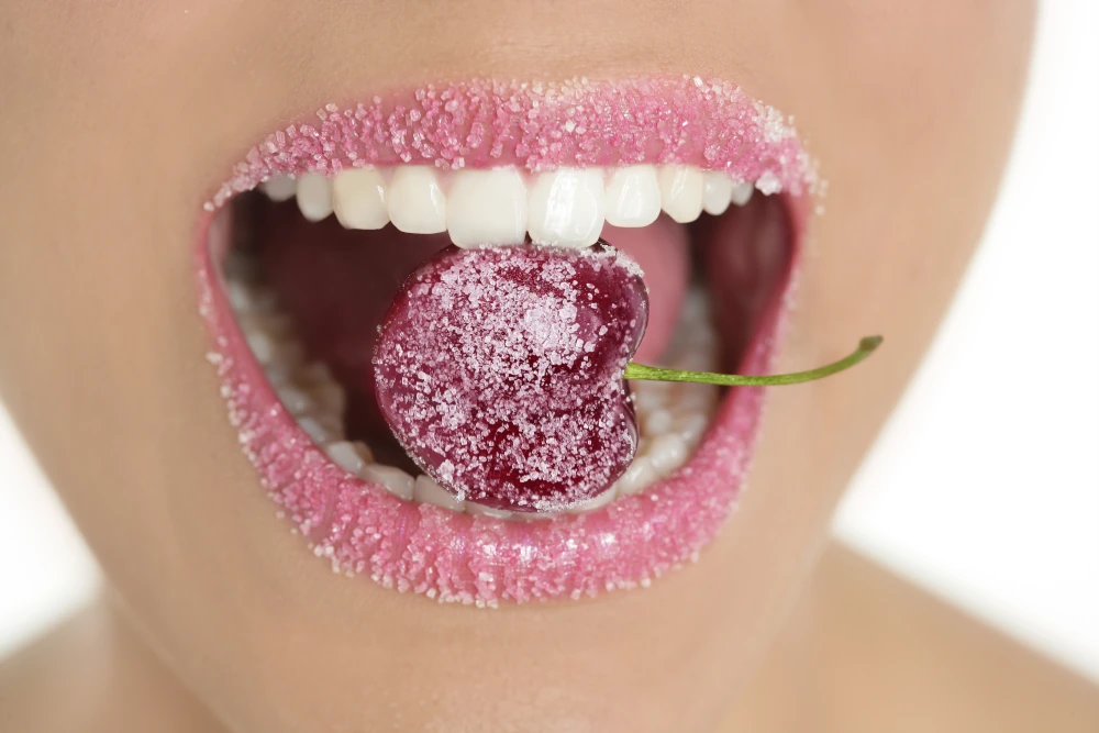 Zu sehen ist der offene Mund einer jungen Frau. Sie hat strahlend weiße Zähne, rosanen Lippenstift auf den Lippen und zwischen den Zähnen eine mit Zucker bedeckte Kirsche. Auch Ihre Lippen sind komplett voll Zucker. 