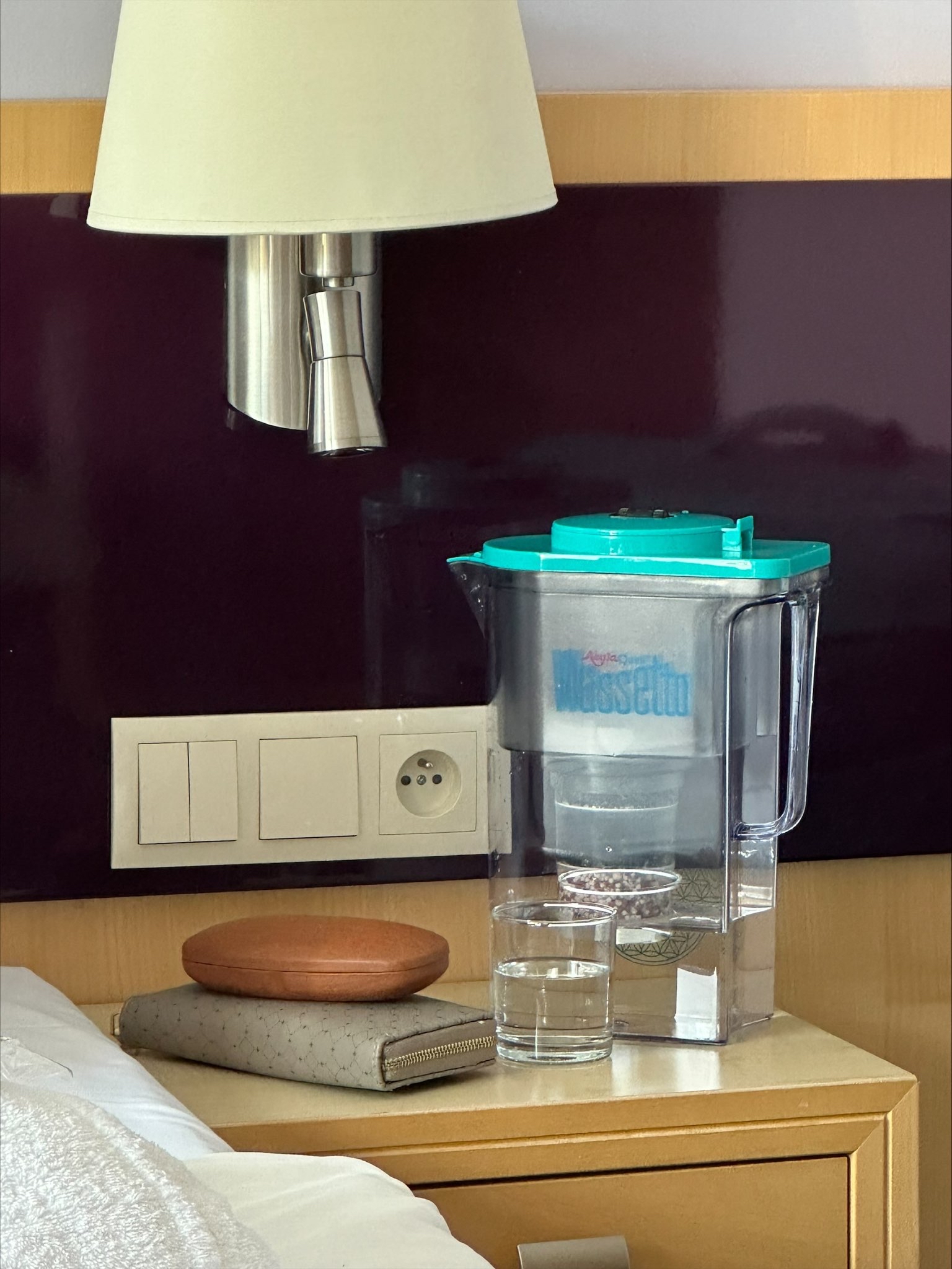Nachttisch auf der rechten Bettseite in einem Hotelzimmer.Auf dem Tisch liegt eine Geldbörse,ein Brillenetui und ein Kannenwasserfilter Wassetto