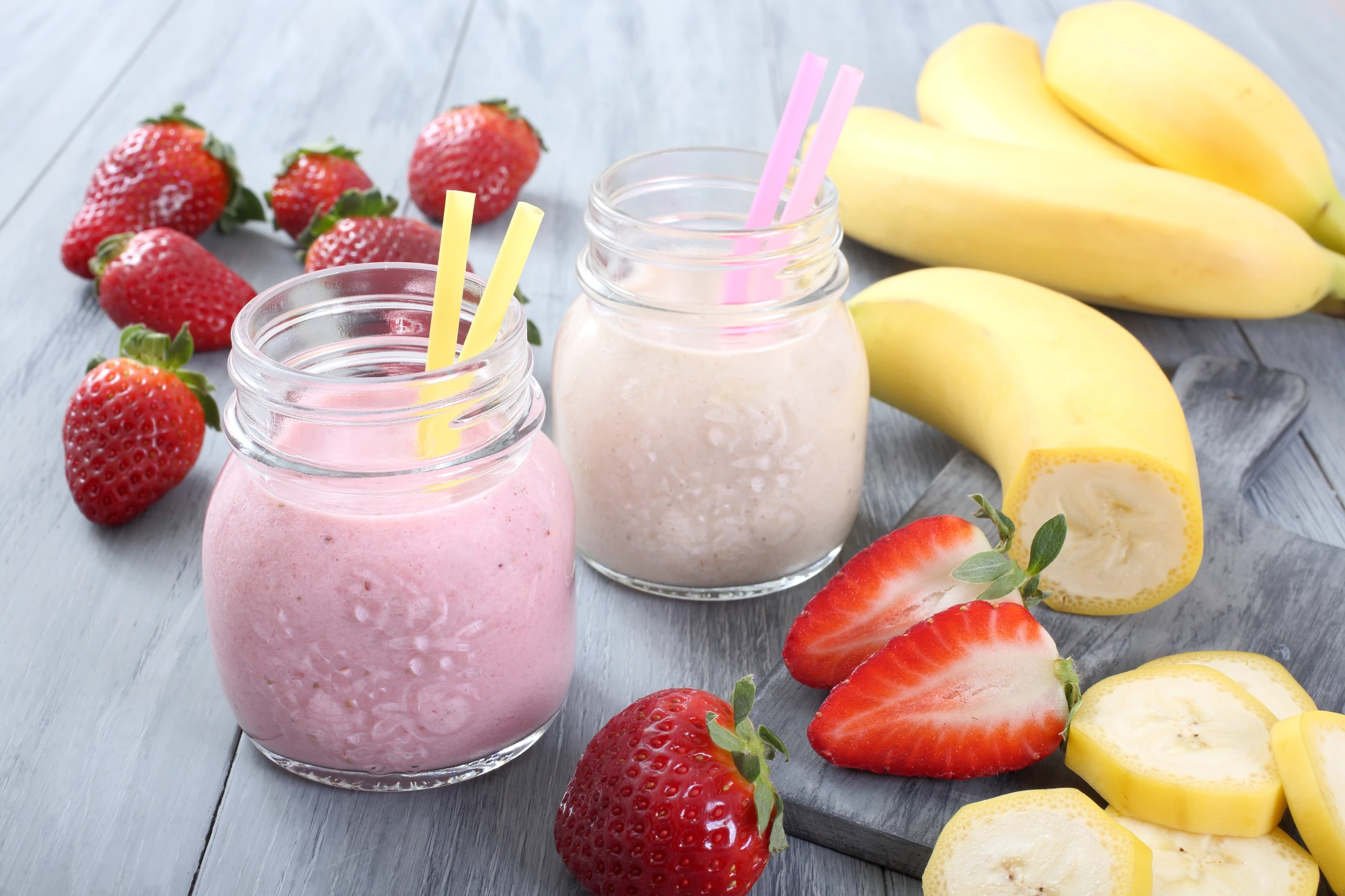 Zu sehen sind zwei Gläser mit Smoothies oder Jogurt darin. In jedem Glas sind zwei Strohhalme, um die Gläser herum liegen frische Erdbeeren und Bananen. 