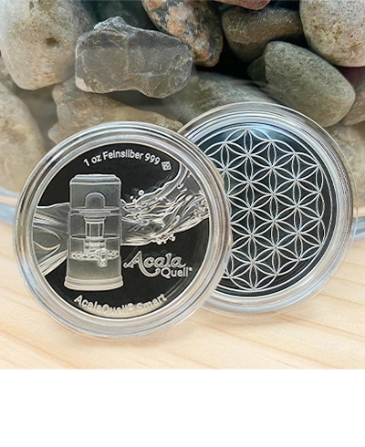 Zu sehen ist eine Acala Silbermünze auf einer hellen Holzplatte vor einem Glas, in dem verschiedene Steine sind in Großaufnahme. Die Münze ist von Vorder- und Rückseite zu sehen. Vorne ist der AcalaQuell Smart, hinten die Blume des Lebens.