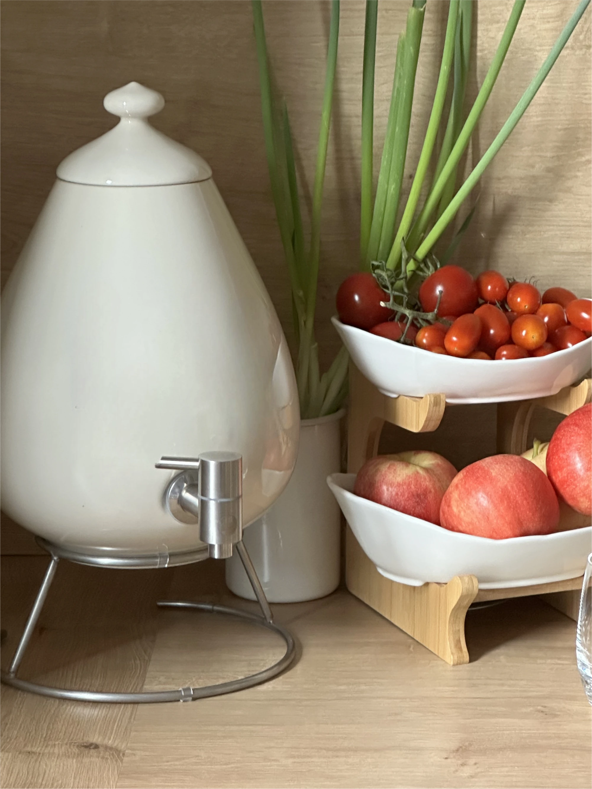 AcalaQuell® Porzellan-Ei Man sieht ein Porzellan Ei auf einem Metallständer und Edelstahl Wasserhahn auf einer Küchenarbeitsplatte. Daneben eine Schale mit frischem Obst und Gemüse. Das Bild zeigt das Porzellan Ei 5L in beige aus Handarbeit von Acala.