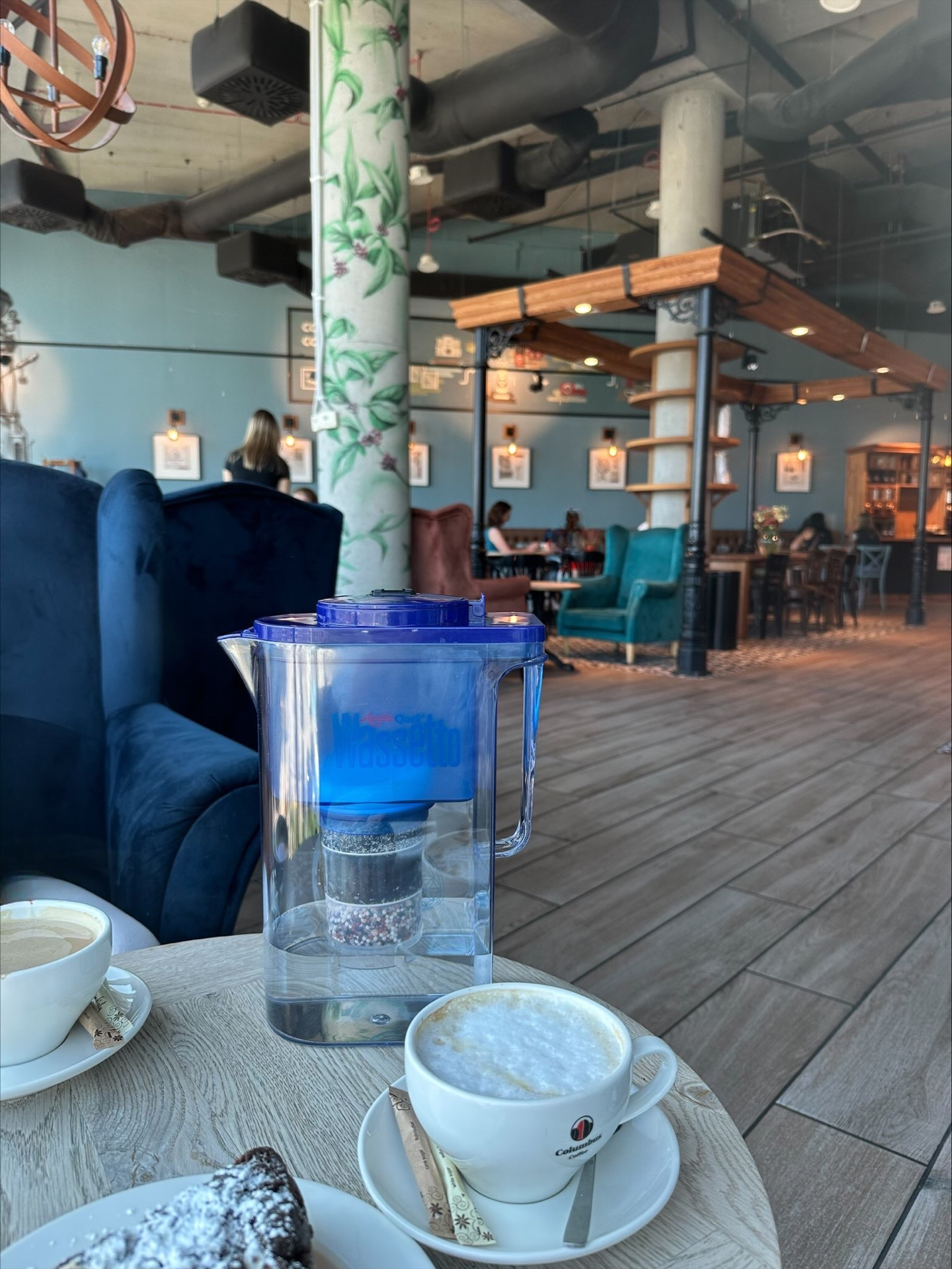 Wassetto Kannen Wasserfilter, 2 Kaffee Tassen und ein Teller mit Kuchen auf einem kleinen Holztisch. Im Hintergrund sieht man ein Cafe im industriellen Stil. 