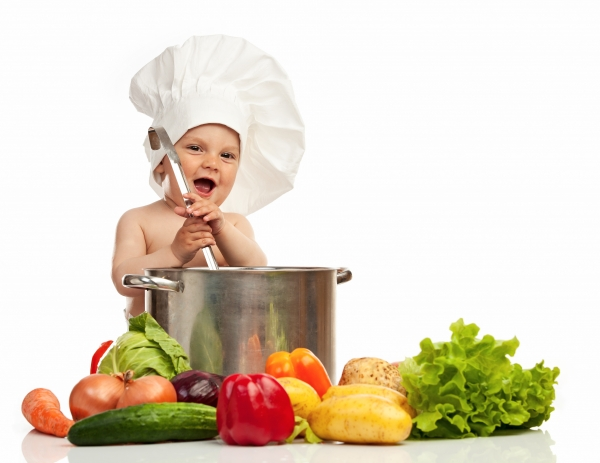 Zu sehen ist ein großer Kochtopf vor dem viel verschiedenes Gemüse liegt. Kartoffeln, Gurken, Zwiebeln, Paprika und Salat. Hinter dem Topf sieht man ein lachendes Kleinkind mit großer Kochmütze auf dem Kopf, welches mit einer Kelle im Topf rührt. 
