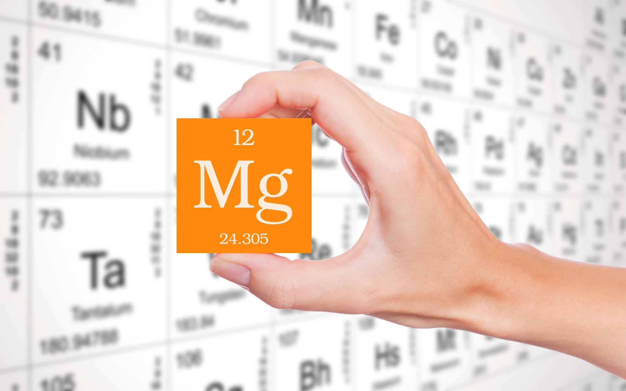 Eine Hand hält einen orangenen Würfel in der Hand auf dem Mg steht. Im Hintergrund ist das Periodensystem zu sehen.