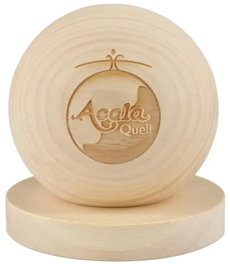 Zu sehen ist eine helle Holzkugel die auf einem hellen Holzteller liegt vor weißem Hintergrund. Auf der Kugel sieht man das Acala Logo. Das Bild zeigt die Zirbenholzkugel mit Ablageteller von Acala.