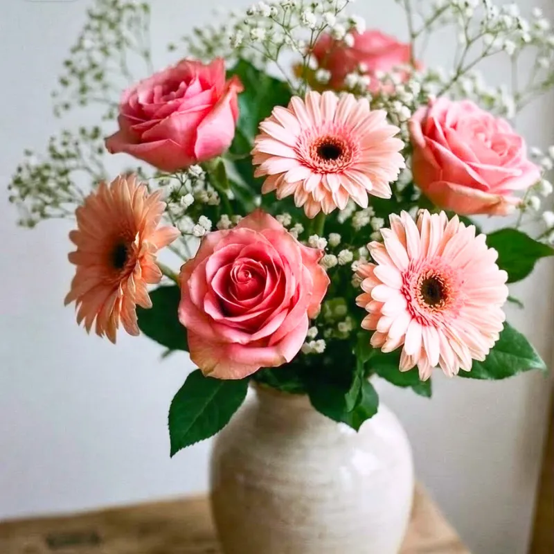 Ein Strauß mit hellrosa Germerer weißem Schleierkaut und pinken Rosen in einer beigen Steinvase