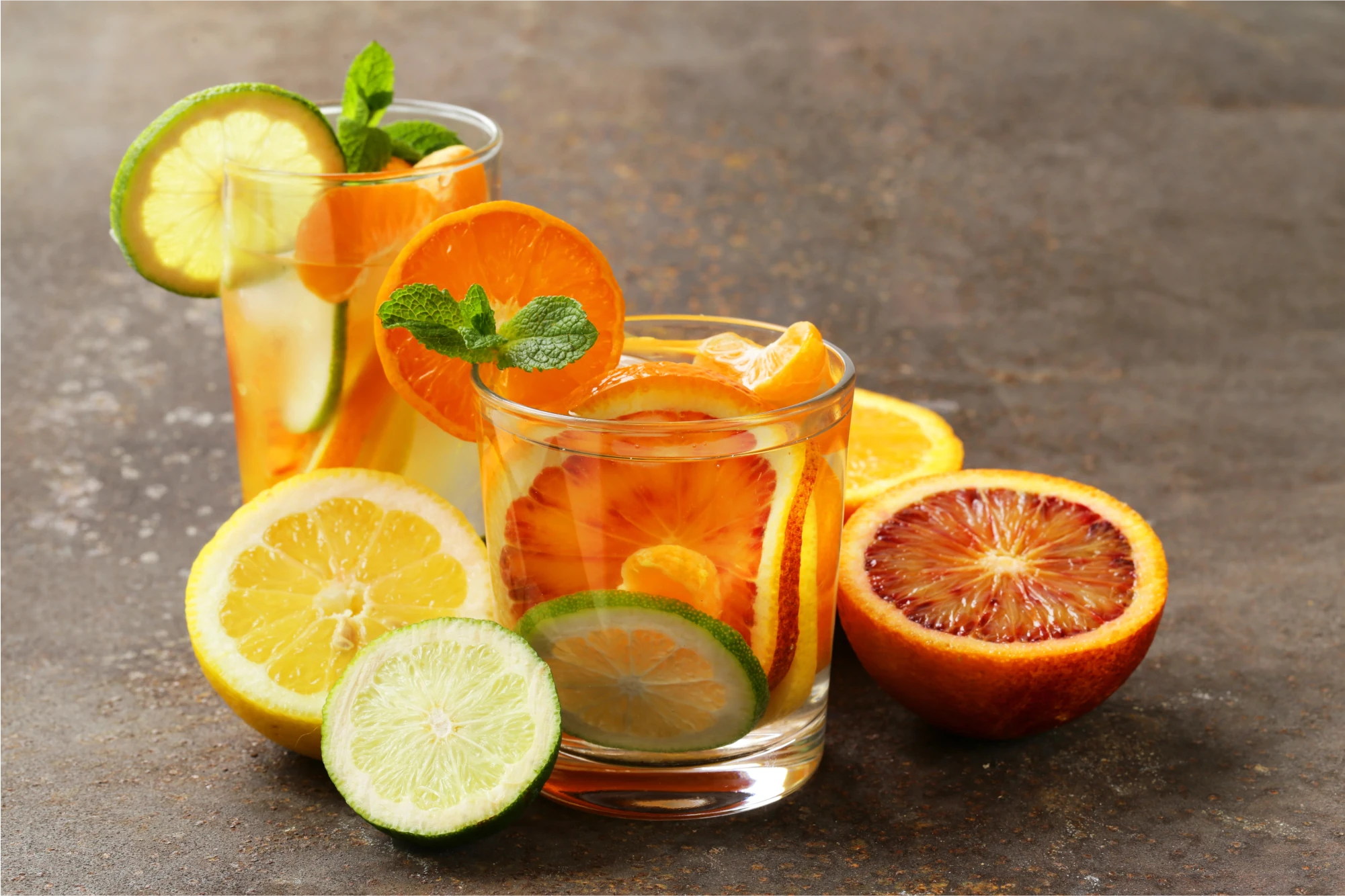 Zwei Gläser mit einem Getränk, das mit Scheiben von Zitrone, Limette, Orange und Blutorange sowie Minzblättern garniert ist auf dunklem Untergrund.