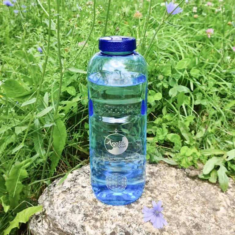 Zu sehen ist eine mit Wasser gefüllte Tritan Trinkflasche in blau mit blauem Schraubdeckel. Sie steht auf einem Stein, im Hintergrund sieht man eine Wiese, die Sonne scheint. Das Acala Logo ist in weiß auf der Flasche zu sehen. 
