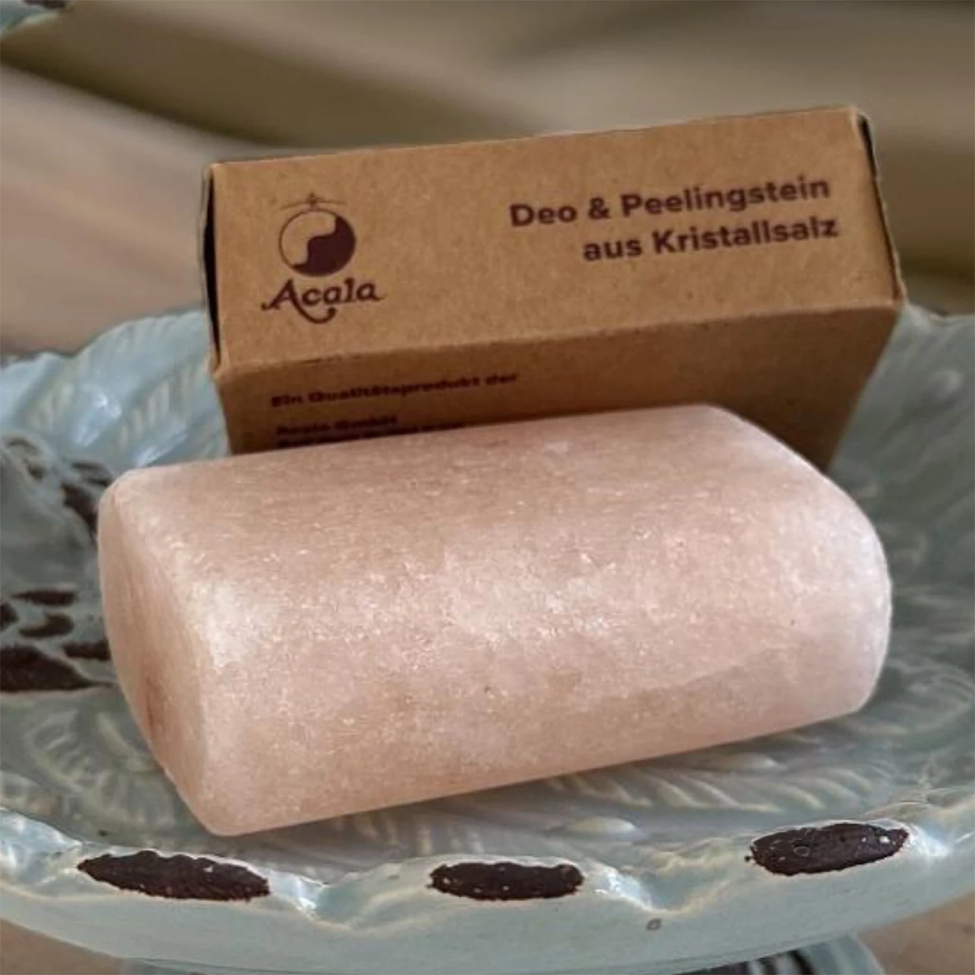Zu sehen ist ein Salz Deo Stein von Acala. Er sieht aus wie ein Stück Seife, liegt in einer Porzellan Schale und dahinter sieht man den braunen Verpackungskarton.
