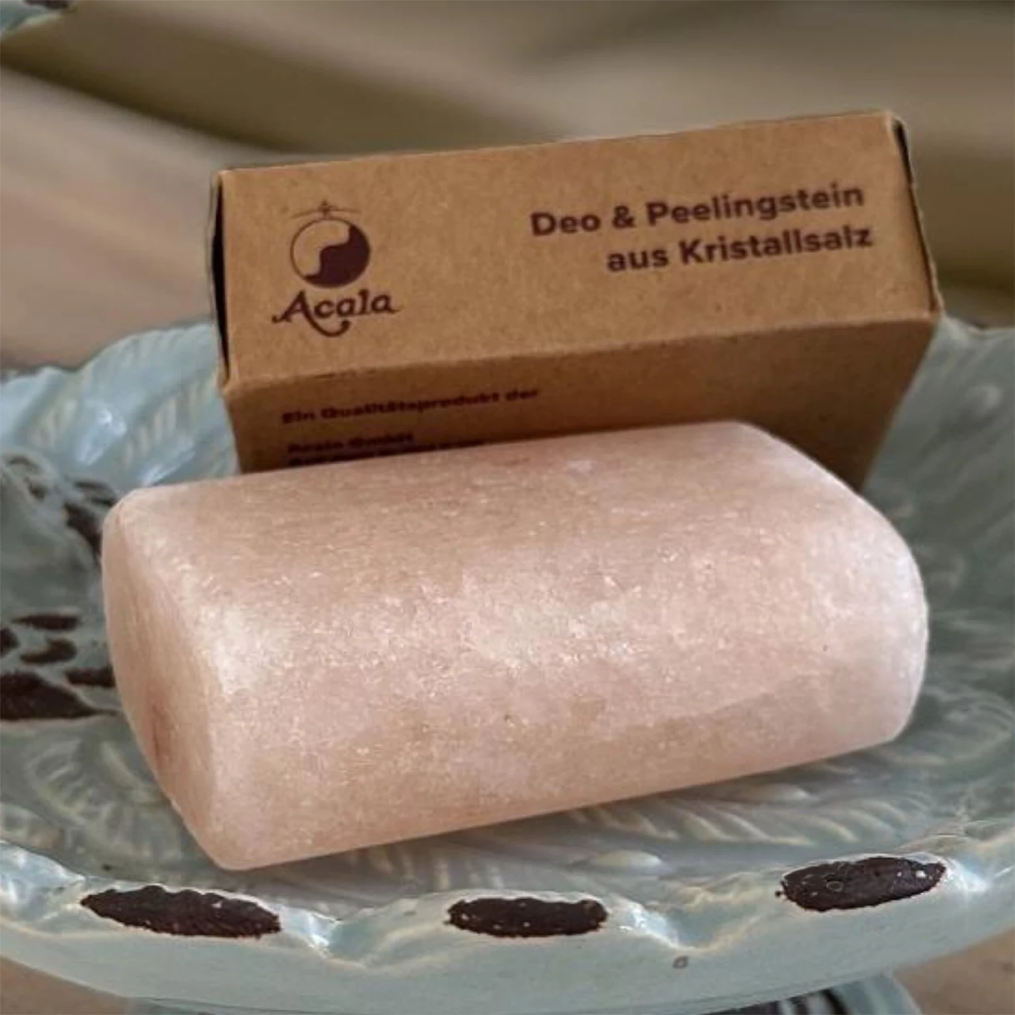 Deo & Peelingstein aus Kristallsalz Zu sehen ist ein Salz Deo Stein von Acala. Er sieht aus wie ein Stück Seife, liegt in einer Porzellan Schale und dahinter sieht man den braunen Verpackungskarton.