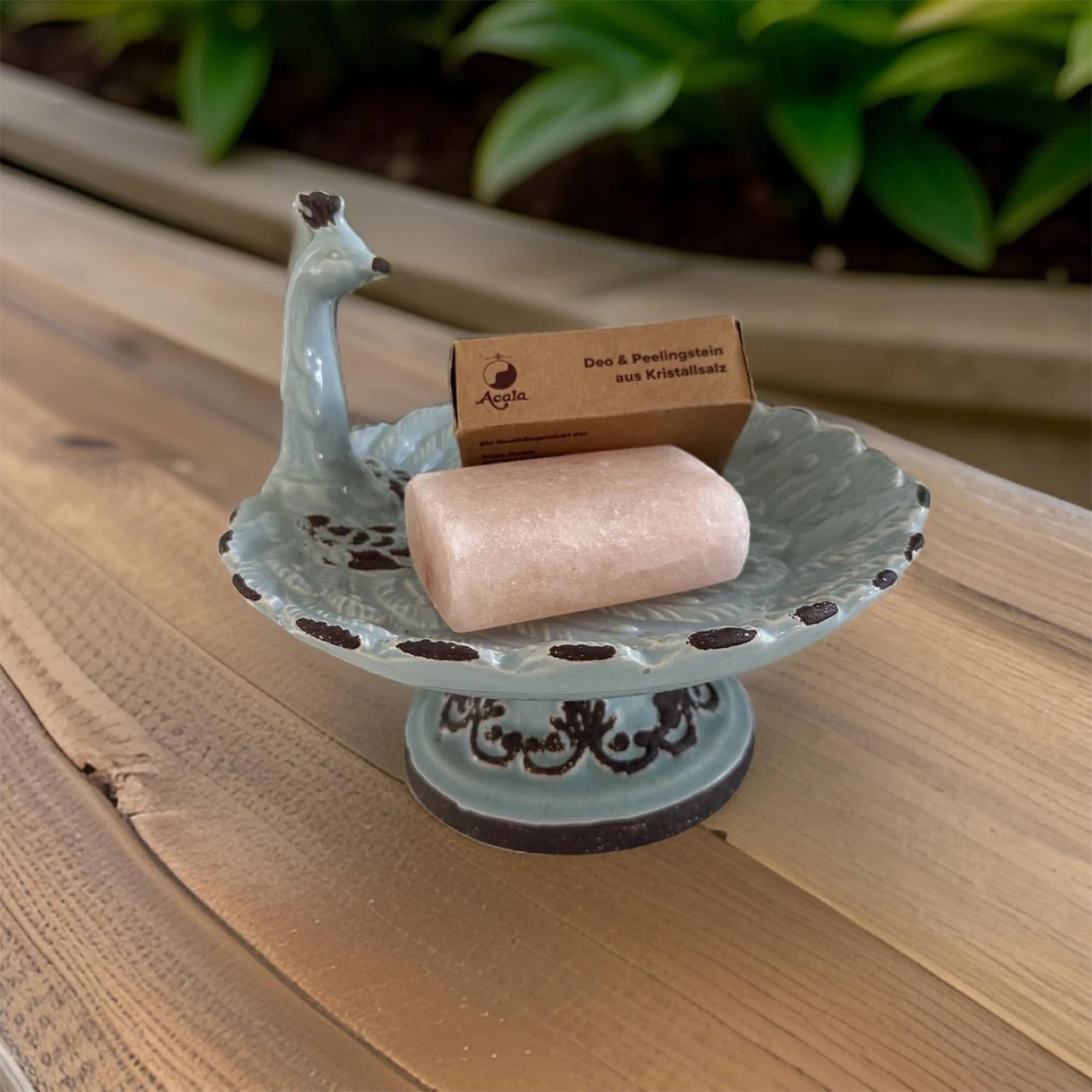 Zu sehen ist ein Stein der aussieht wie ein Stück Seife in hellem rosa. Er liegt zusammen mit dem braunen Verpackungskarton in einer Schale die wie ein Vogel aussieht. Diese steht auf einem braunen Holztisch. Das Bild zeigt den Salz Deo Stein von Acala.