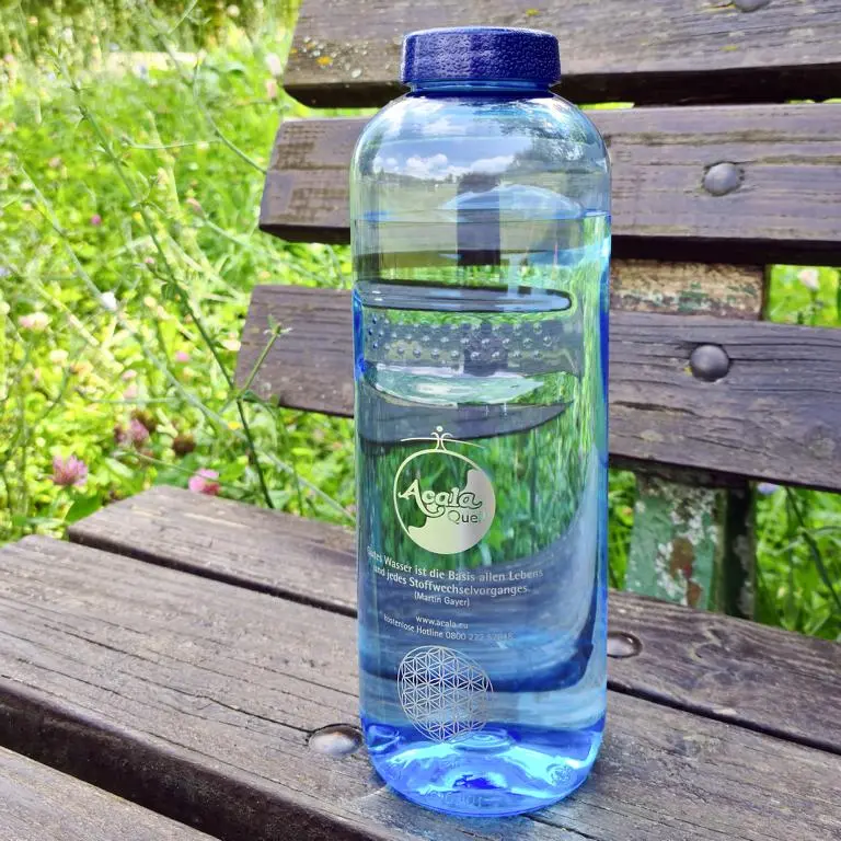 Zu sehen ist eine blaue Tritan Trinkflasche mit dunkelblauem Schraubdeckel. Sie steht auf einer Holzbank, im Hintergrund sieht man eine grüne Wiese. Auf der Flasche sieht man das Acala Logo und die Blume des Lebens.