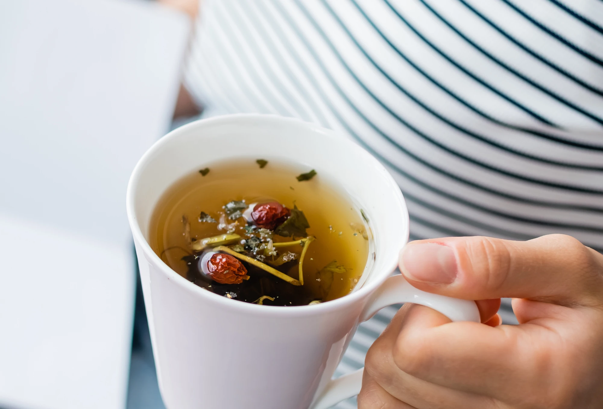 Weibliche Hand hält weiße Tasse mit Tee in der Hand. In dem Tee schwimmen verschiedene Zutaten, wie Kräuter und Beeren.
