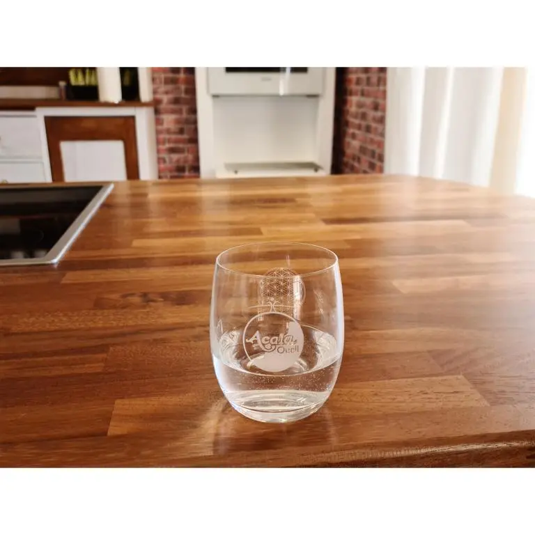 Zu sehen ist ein zur Hälfte mit Wasser gefülltes Trinkglas, Modell Arlando von Acala auf einer dunklen Holzarbeitsplatte. Im Hintergrund sieht man eine Küch