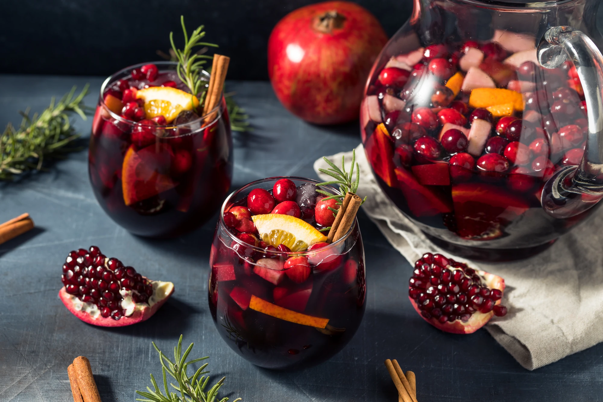 Ein Getränk in zwei Gläsern und einem Krug, gefüllt mit roten Früchten wie Granatapfel, Orangenscheiben, Zimtstangen und Rosmarinzweigen. Es handelt sich um eine festliche, rote Fruchtbowle auf einem dunklen Hintergrund.