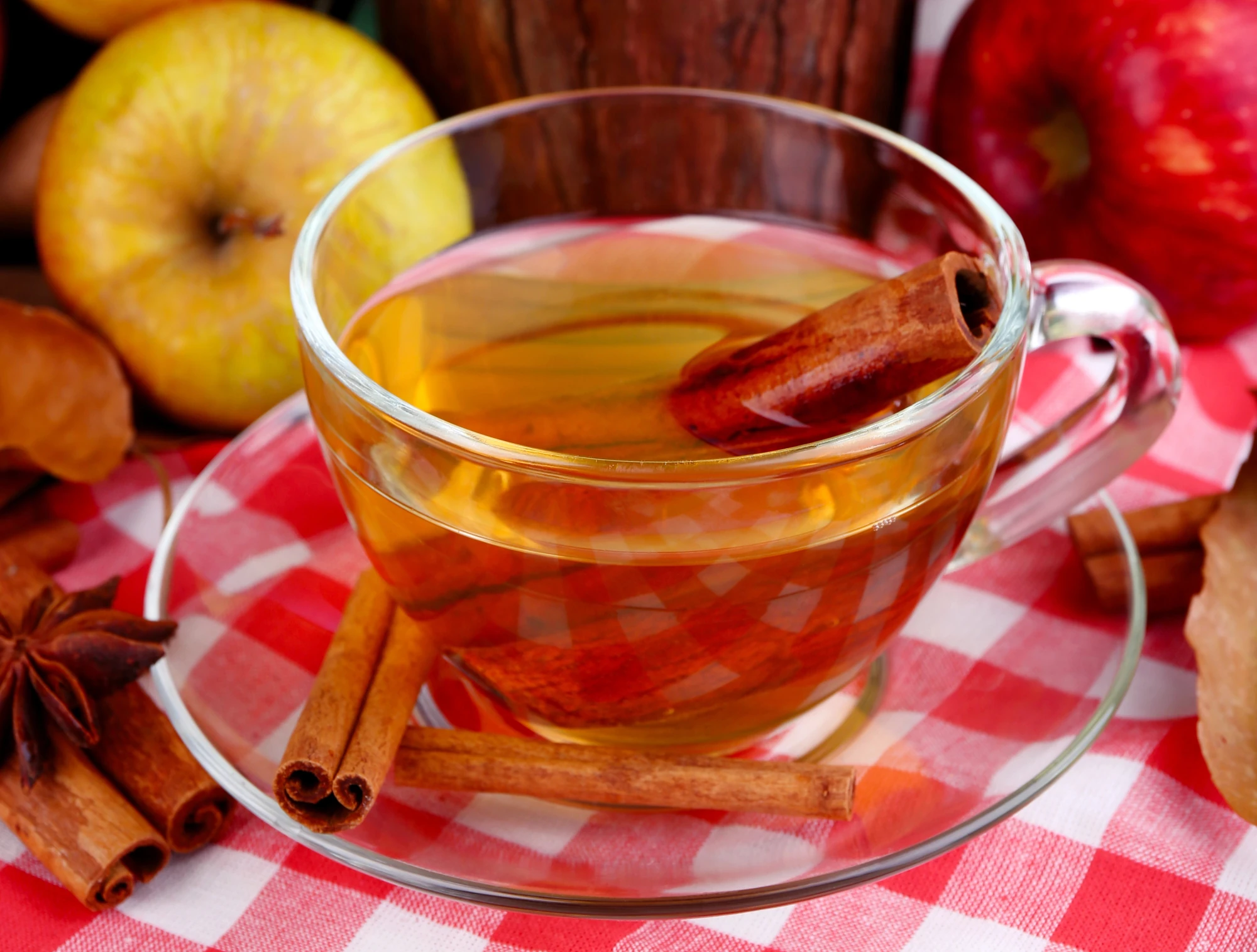 Durchsichtige kleine Tasse mit hell bräunlicher Flüssigkeit auf durchsichtigem Unterteller. Zimtstangen liegen auf dem Teller, eine ist in der Flüssigkeit. Rustikale Atmosphäre.