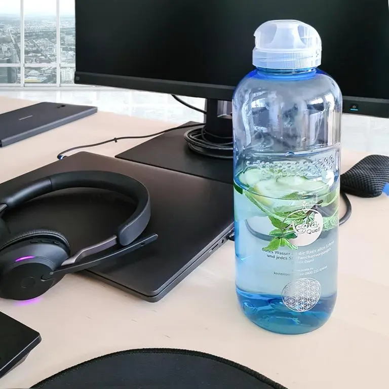 Zu sehen ist eine blaue Tritan Trinkflasche mit transparentem Klappdeckel auf einem Schreibtisch in einem Büro. Die Flasche ist mit Wasser gefüllt, man sieht das Acala Logo und die Blume des Lebens auf der Flasche.