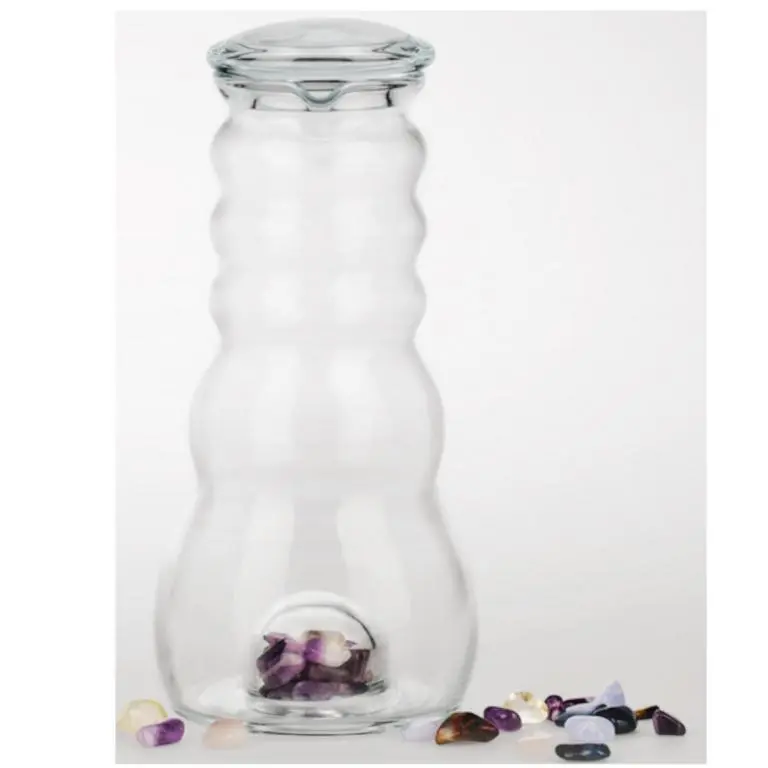 Zu sehen ist eine Glaskaraffe mit Glasdeckel vor weißem Hintergrund. Im unteren Teil hat sie ein separates Fach, in dem sich verschieden farbige Edelsteine befinden. Diese liegen zudem um die Karaffe herum. Das Bild zeigt den Acala Cadus Krug.