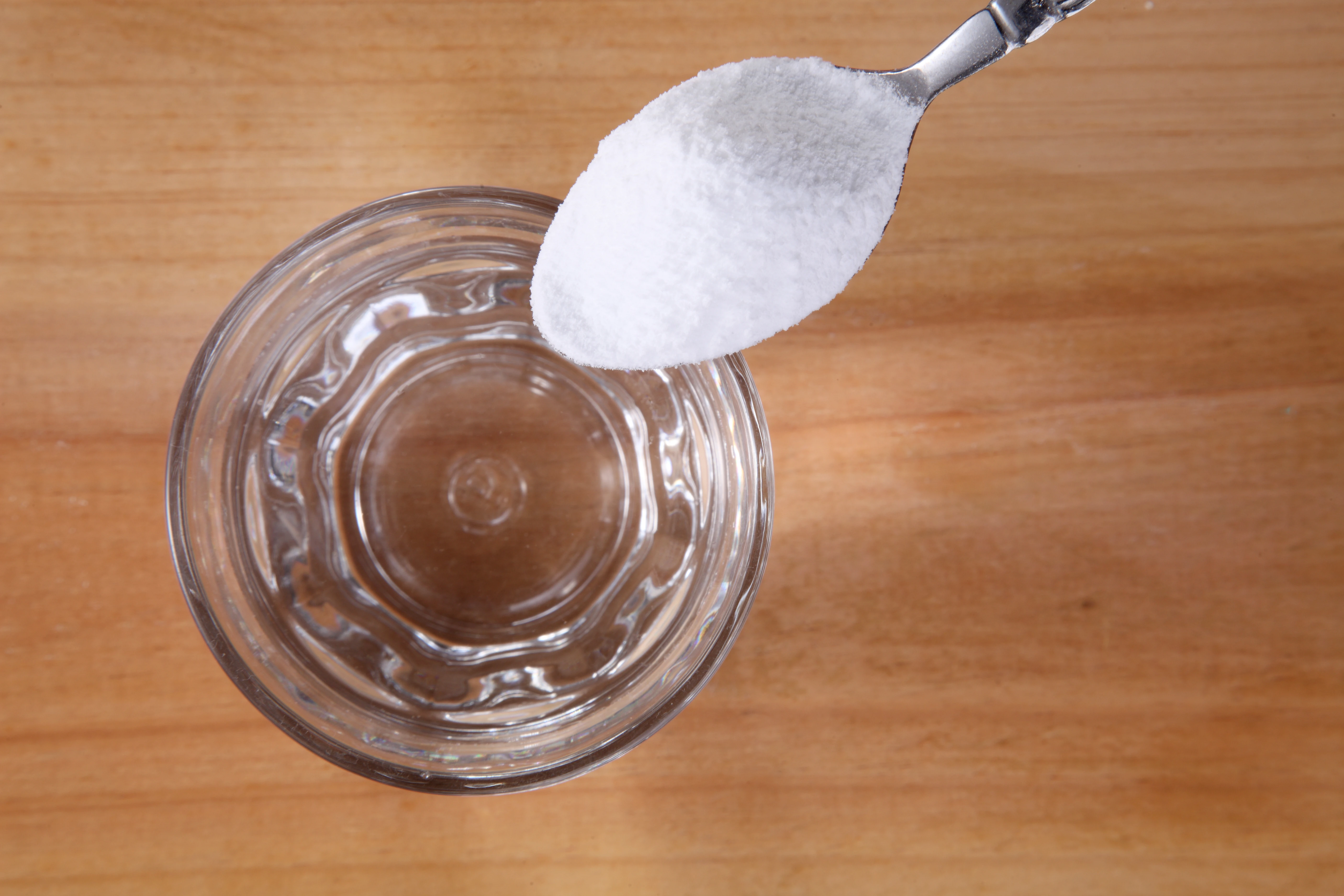 Zu sehen ist ein Glas Wasser, welches auf einer braunen Platt steht. Man schaut von oben drauf und über das Glas hält jemand einen Teelöffel mit weißem Pulver, als wollte er es gleich in das Glas schütten.