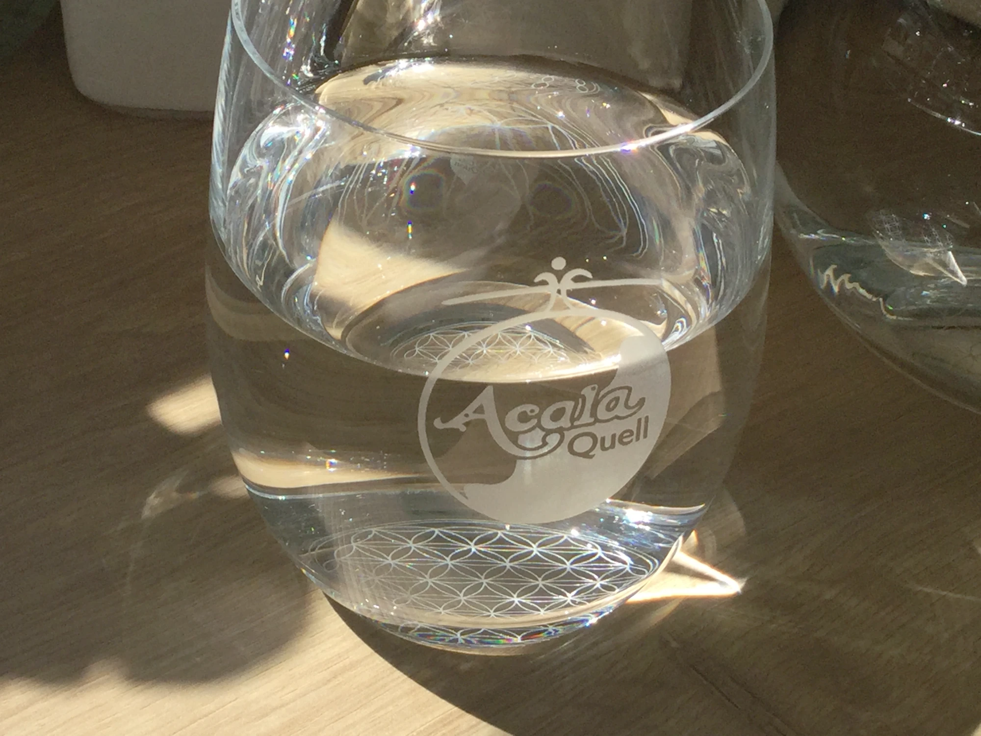Zu sehen ist ein Acala Trinkglas Arlando mit dem AcalaQuell Logo und der Gravur der Blume des Lebens auf dem Boden. Es ist mit Wasser gefüllt und steht auf einem Tisch. Sie Sonne, die durch ein Fenster einfällt spiegelt sich im Glas und im Wasser.