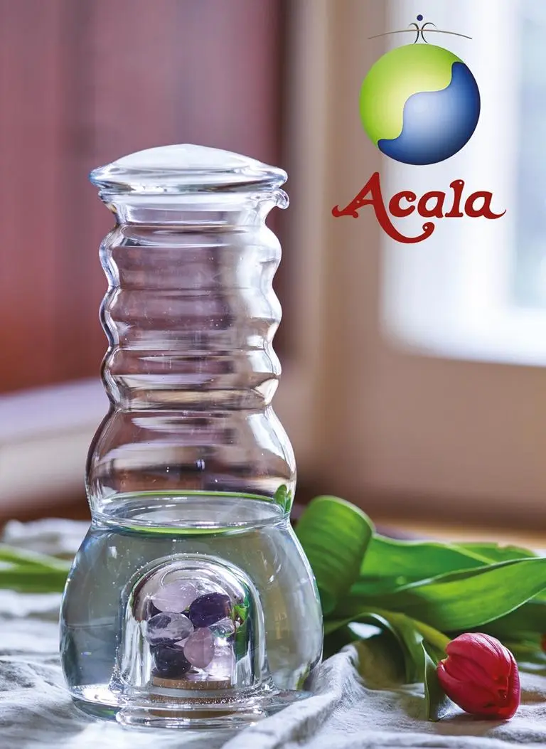 Cadus Krug 1.5 Liter mit Edelsteinfach Zu sehen ist eine Glaskaraffe mit flachem Glasdeckel. Sie steht auf einem Tisch, hinter ihr liegt eine rote Tulpe. Im unteren Bereich der Karaffe sieht man ein Fach, in dem sich verschiedene Edelsteine befinden. Das Bild zeigt den Cadus Krug von Acala.