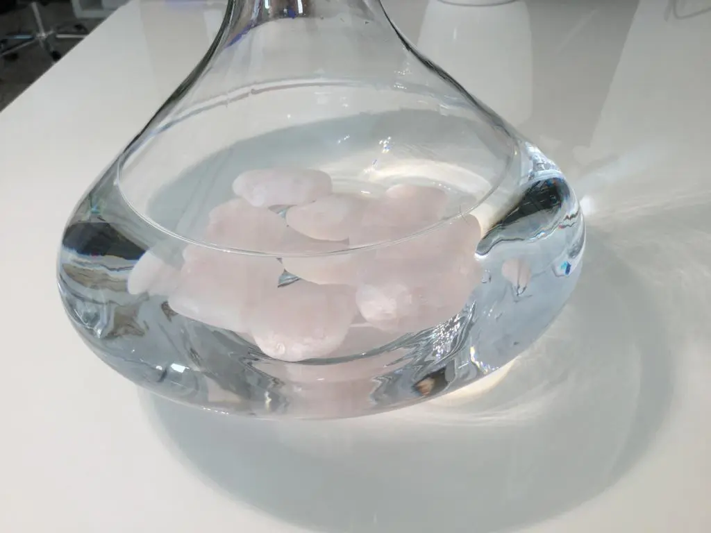 Zu sehen ist eine mit Wasser gefüllte Glaskaraffe, die auf einem Tisch steht. In der Karaffe liegen mehrere rosane Edelsteine am Boden. Das Bild zeigt die Rosenquarz Wassersteine von Acala.