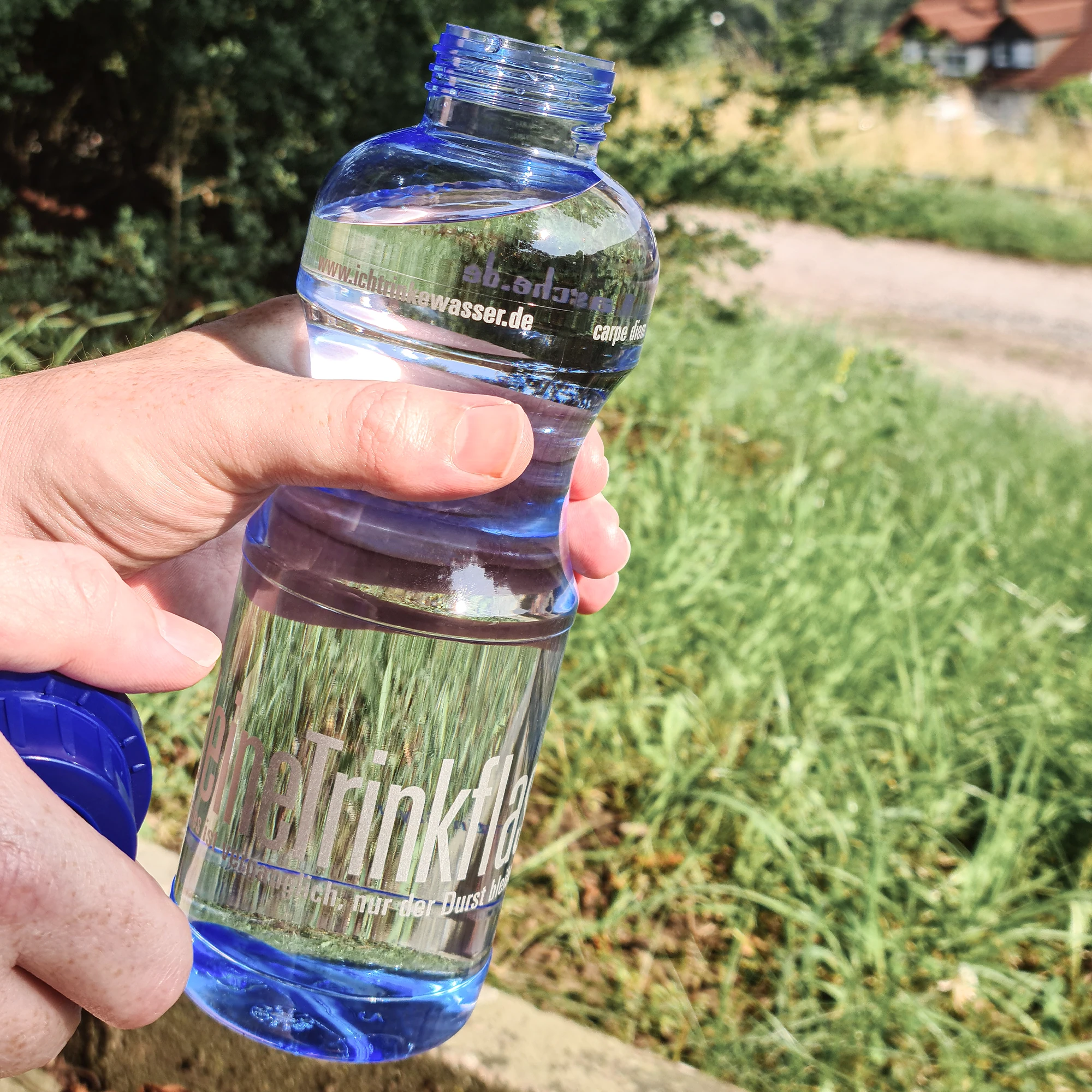 Eine Hand hält eine offene mit Wasser gefüllte blaue Tritan Trinkflasche Adam 0,75L. Im Hintergrund eine grüne Wiese.