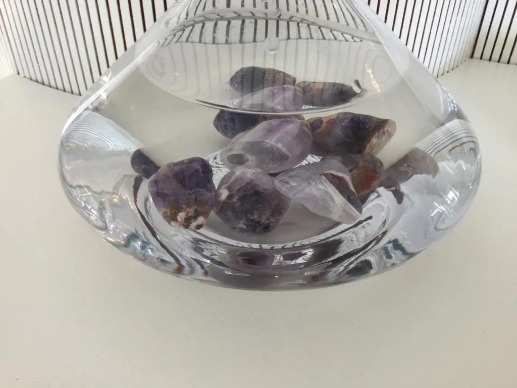 Zu sehen ist der untere Teil einer mit Wasser gefüllten Glaskaraffe, die auf einem Tisch steht. In der Karaffe liegen mehrere lila farbene Edelsteine am Boden. Das Bild zeigt die Amethyst Wassersteine von Acala.