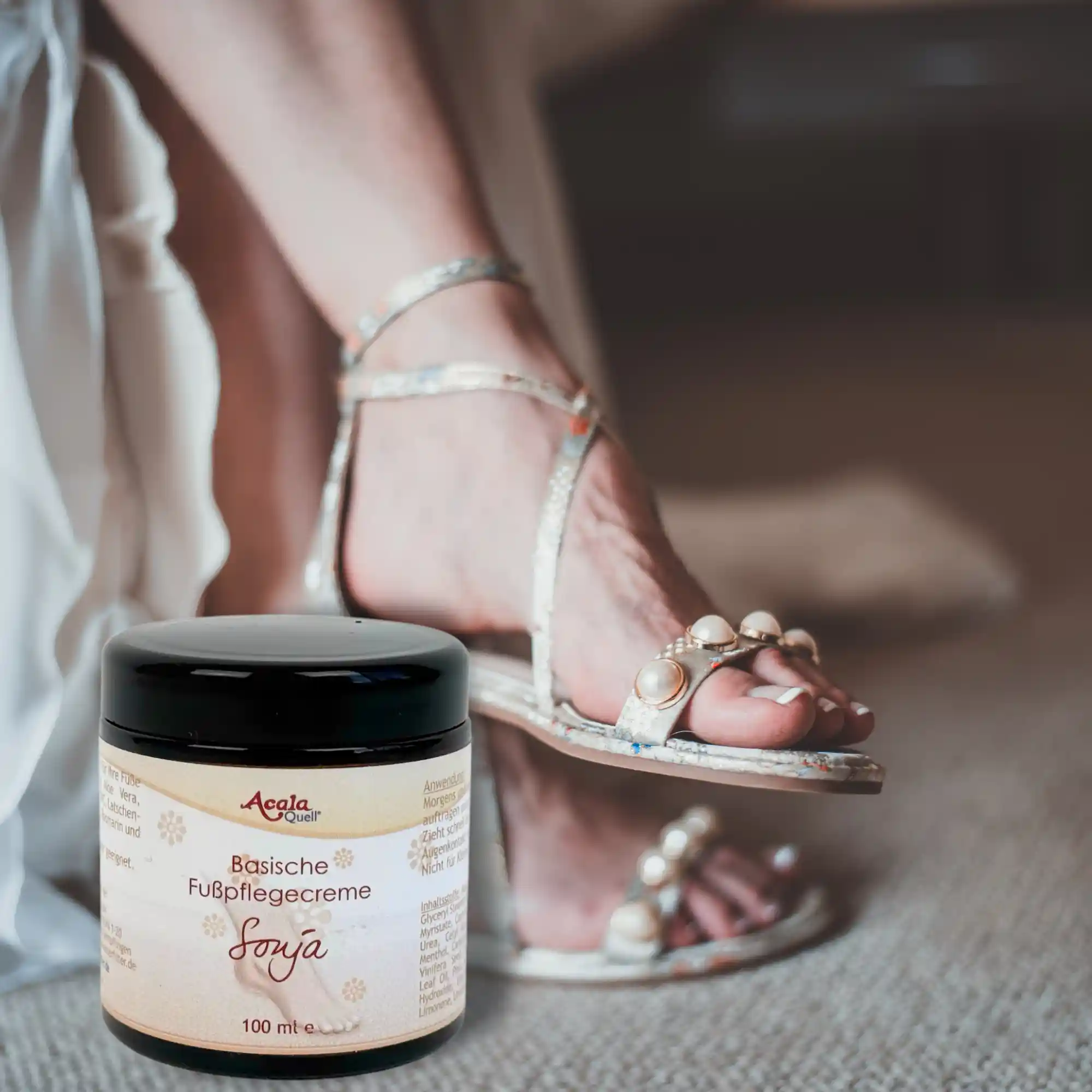Füsse in Sandalen im Hintergrund, im Vordergrund ein Cremtigel aus braunem Gls mit dem Etikett der Sonja Fusspflege