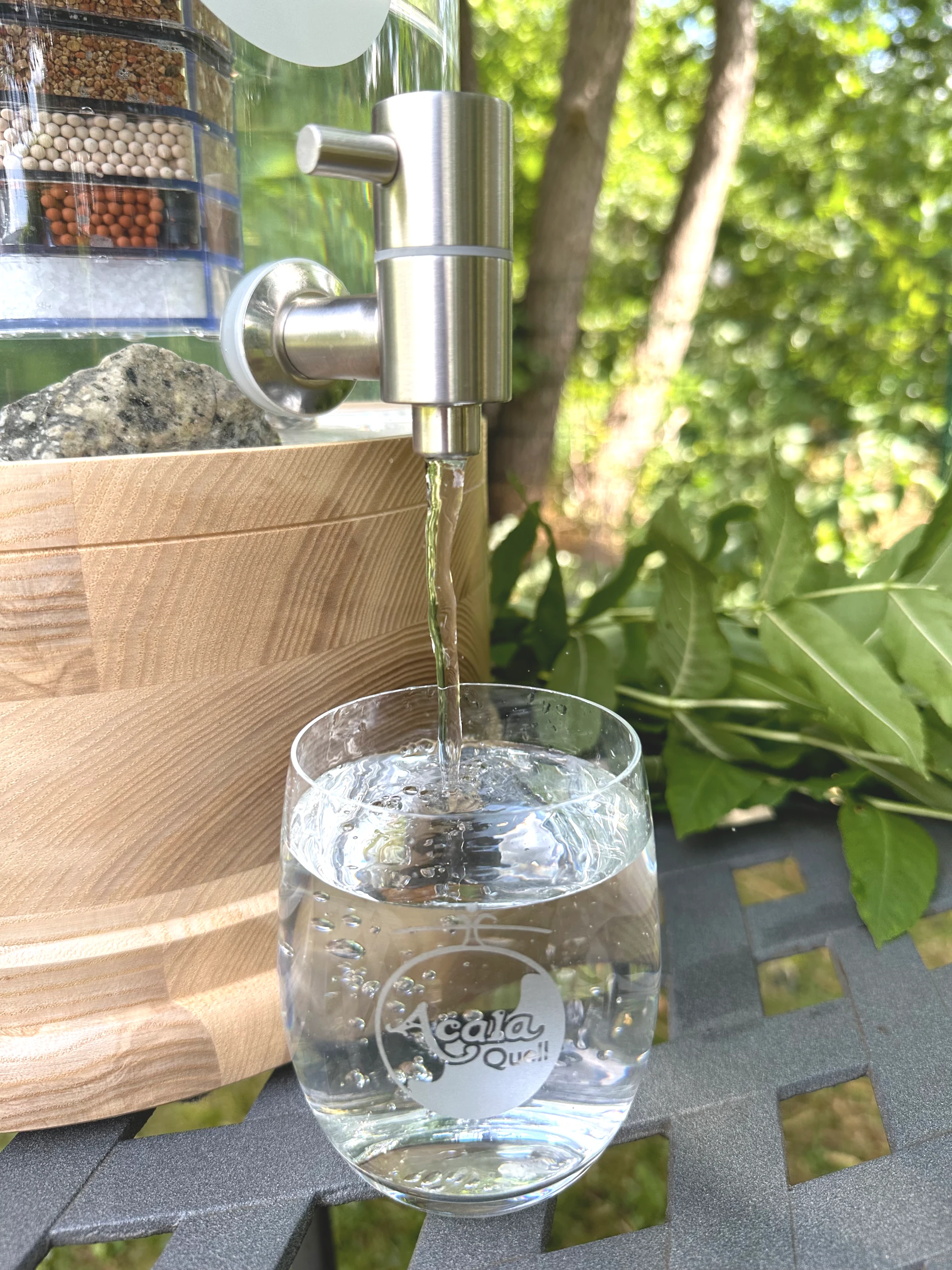 Zu sehen ist ein Ausschnitt eines Standwasserfiltes mit Echtholzsockel aus Esche. Aus dem Edelstahl Wasserhahn läuft Wasser in ein darunter stehendes Glas. Um den Filter herum sieht man grüne Blätter und Bäume.