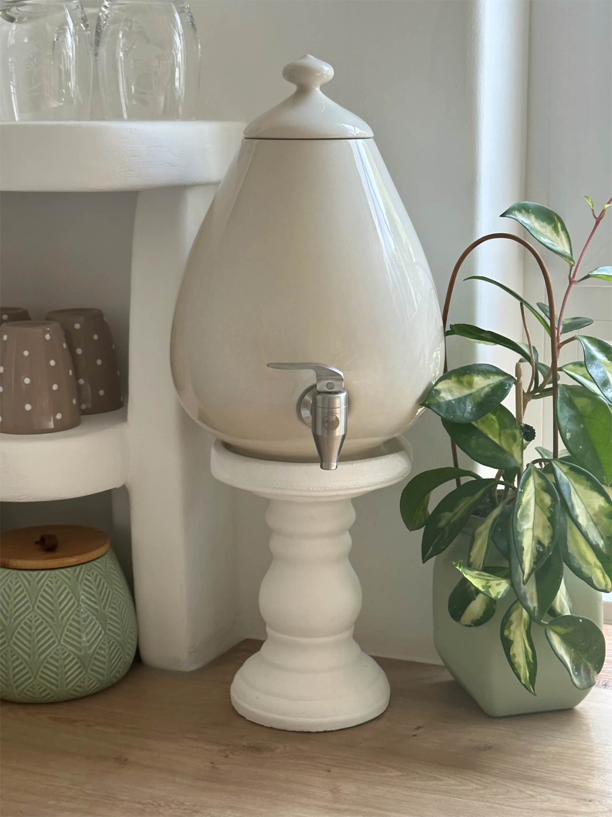 Zu sehen ist ein 5L Porzellan Ei aus Handarbeit von Acala. Es steht auf einem weißen Sockel. Es ist beige und es ist Edelstahl Wasserhahn daran. Neben dem Ei sieht man ein Regal mit Tassen und eine grüne Topfpflanze.