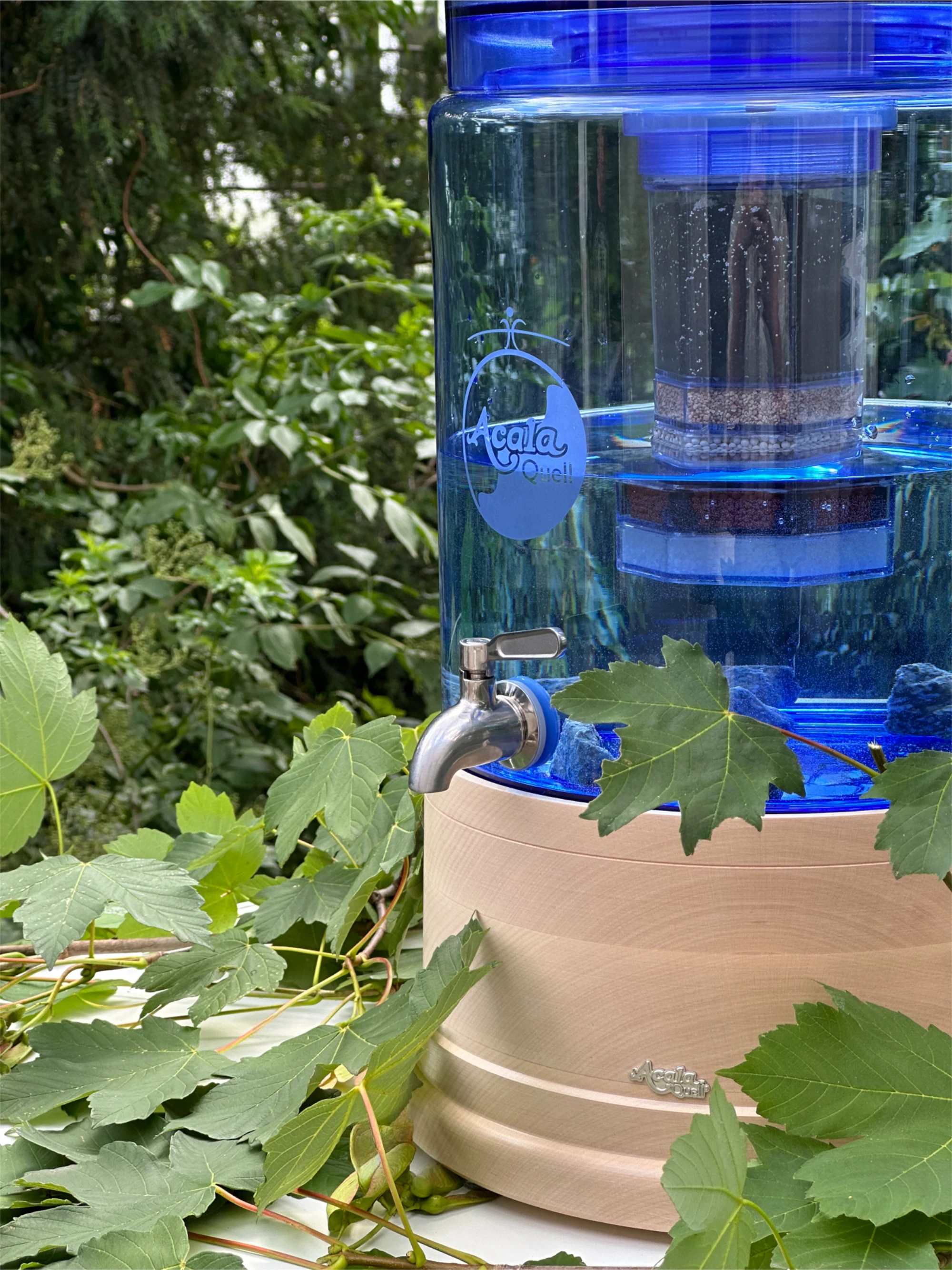 Zu sehen ist ein Standwasserfilter mit Glastank in blau und Ahorn Echtholz Sockel und Deckel. Der Filter steht auf einem weißen Tisch, im Hintergrund sieht man einen grünen Baum, um den Filter herum liegen grüne Blätter. 