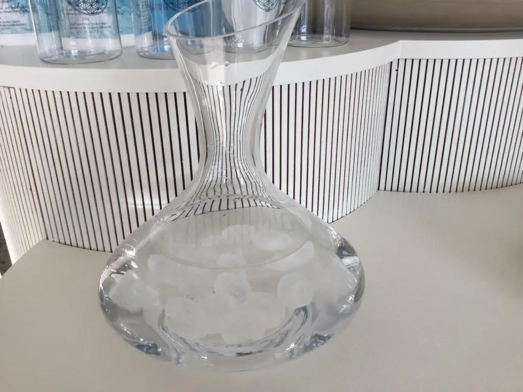 Eine mit Wasser gefüllte Glaskaraffe auf einem weißen Tisch. In der Karaffe befinden sich weiße Bergkristall Edelsteine.