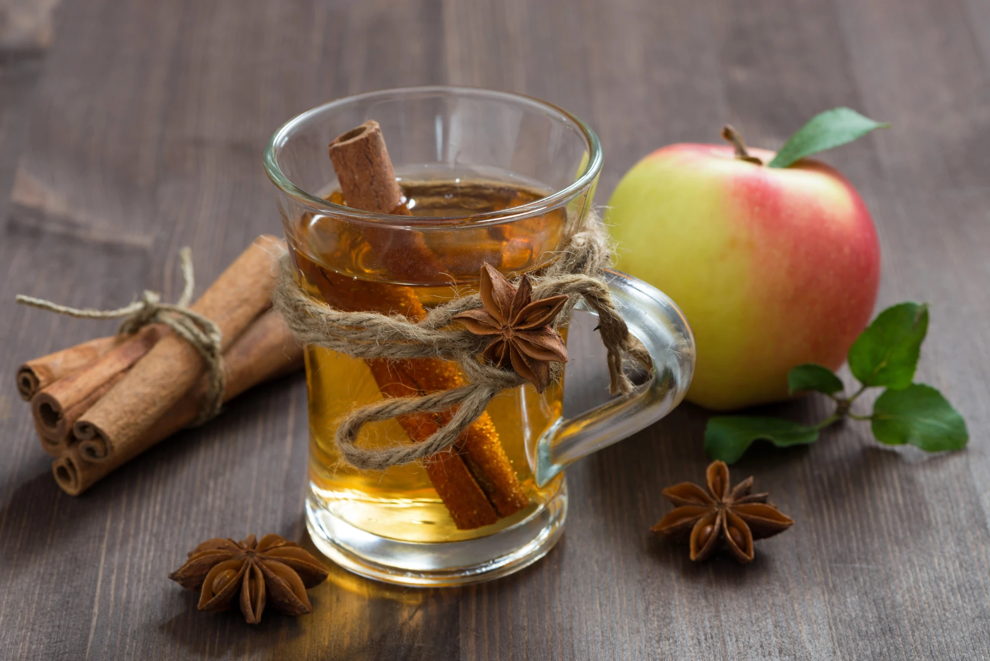 Durchsichtiges Glas mit Henkel mit bräunlicher Flüssigkeit darin auf dunklem Holztisch. In der Flüssigkeit steckt eine Zimtstange, neben dem Glas liegt ein Apfel und Zimtstangen.